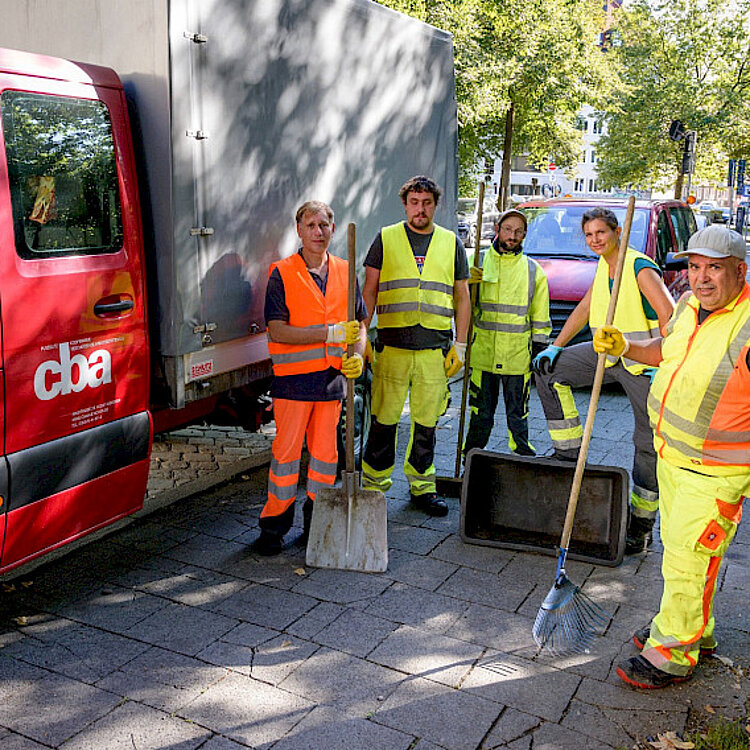 Fünf Straßenarbeiter in neongelber und oranger Warnkleidung stehen auf einem Bürgersteig neben einem roten Fahrzeug. Sie halten Besen und Schaufeln und blicken in die Kamera. Im Hintergrund sind Bäume und geparkte Fahrräder zu sehen.