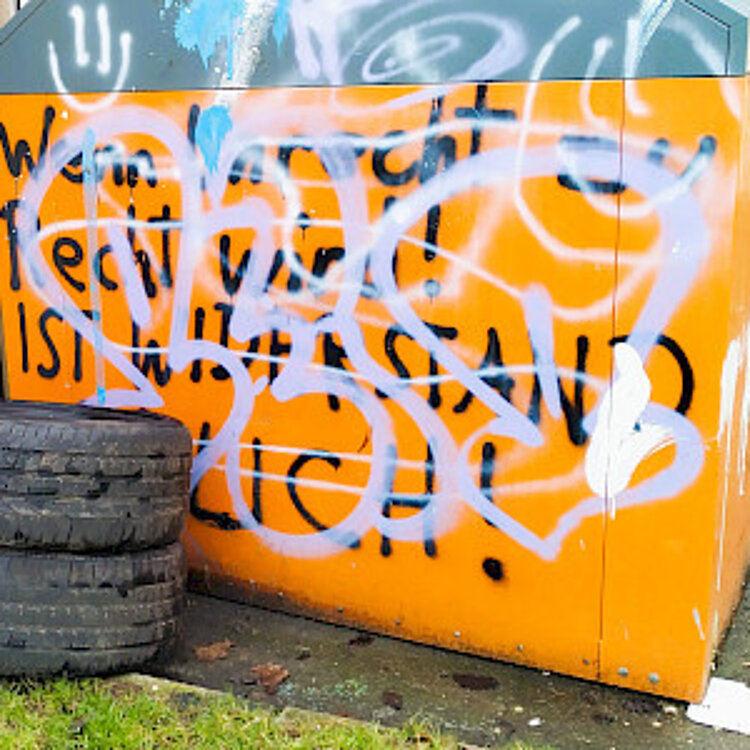 Ein orangefarbener Müllcontainer mit unleserlicher weißer und schwarzer Graffiti-Beschriftung. Daneben liegen drei alte Autoreifen auf dem Boden.
