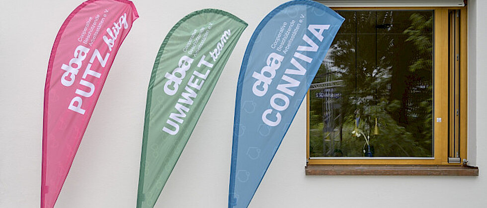 Drei bunte Werbefahnen vor einer weißen Wand mit Fenster, beschriftet mit „PUTZ blitz“, „UMWELT team“ und „CONVIVA“ in großen Buchstaben.