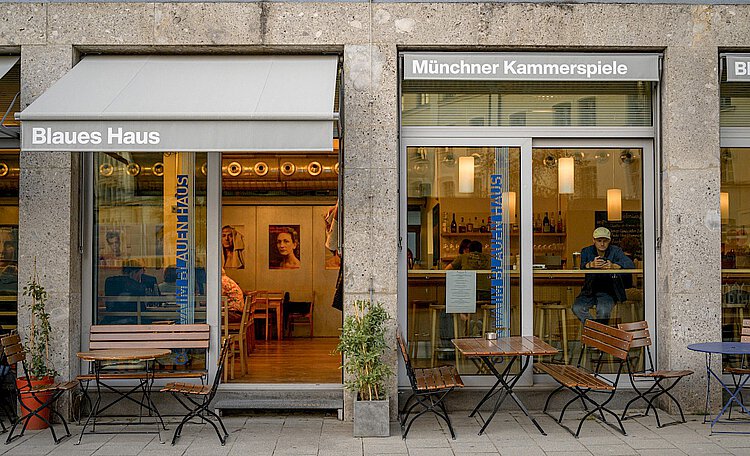 Eingangstür Conviva Außenansicht des Cafés und Kulturlokals „Blaues Haus“ neben den Münchner Kammerspielen mit Tischen und Stühlen vor dem Gebäude.