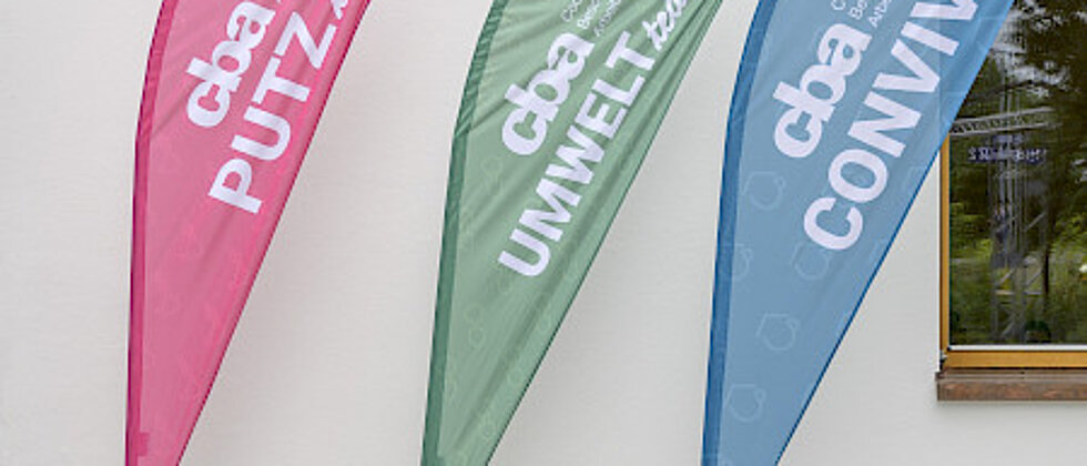 Drei bunte Werbefahnen lehnen an einer weißen Wand, beschriftet mit „cba PUTZ flink“ (pink), „cba UMWELT team“ (grün) und „cba CONVIVA“ (blau).