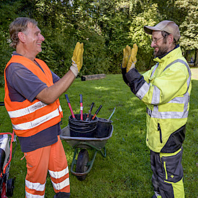 Zwei Bauarbeiter mit Warnkleidung geben sich draußen auf einer Wiese lachend und freudig einen High-Five. Im Hintergrund sind Werkzeuge und ein Schubkarren zu sehen.