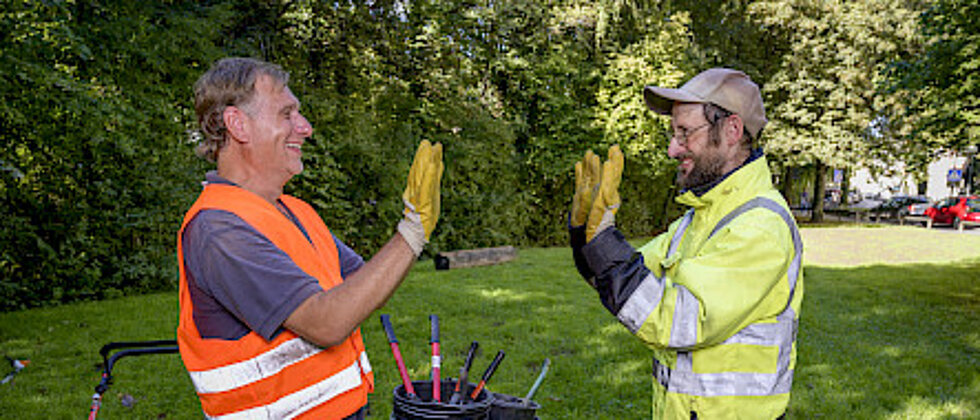 Zwei Bauarbeiter mit Warnkleidung geben sich draußen auf einer Wiese lachend und freudig einen High-Five. Im Hintergrund sind Werkzeuge und ein Schubkarren zu sehen.