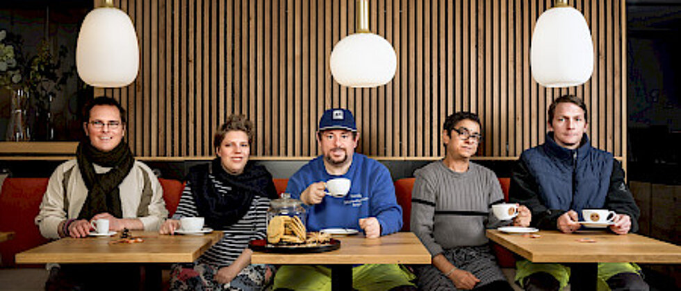 Fünf Personen unterschiedlichen Alters und Geschlechts sitzen an zwei nebeneinanderstehenden Tischen und trinken Kaffee. Sie blicken in die Kamera und wirken entspannt in einem modern eingerichteten Raum mit Holzvertäfelung.