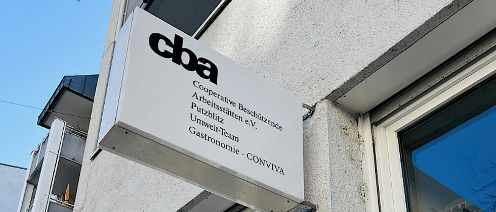 Außenaufnahme eines Gebäudes mit einem Schild, auf dem „cba Cooperative Beschäftigende Arbeitsstätten e.V. Publizistik Umwelt-Team Gastronomie CONVIVA“ steht.