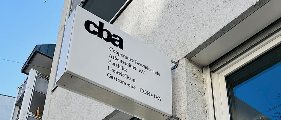 Außenaufnahme eines Gebäudes mit einem Schild, auf dem „cba Cooperative Beschäftigende Arbeitsstätten e.V. Publizistik Umwelt-Team Gastronomie CONVIVA“ steht.
