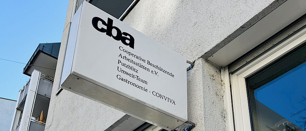 Außenaufnahme eines Gebäudes mit einem Schild, auf dem „cba Cooperative Beschäftigende Arbeitsstätten e.V. Publizistik Umwelt-Team Gastronomie CONVIVA“ steht.