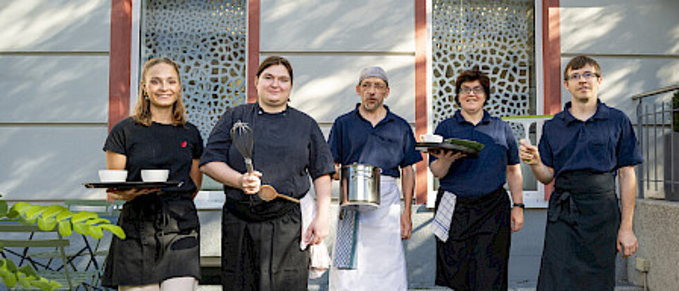 Fünf Personen in Kochkleidung stehen nebeneinander und halten Kochutensilien wie Schneebesen, Topf, Teller und Glas, symbolisieren ein Küchenteam.
