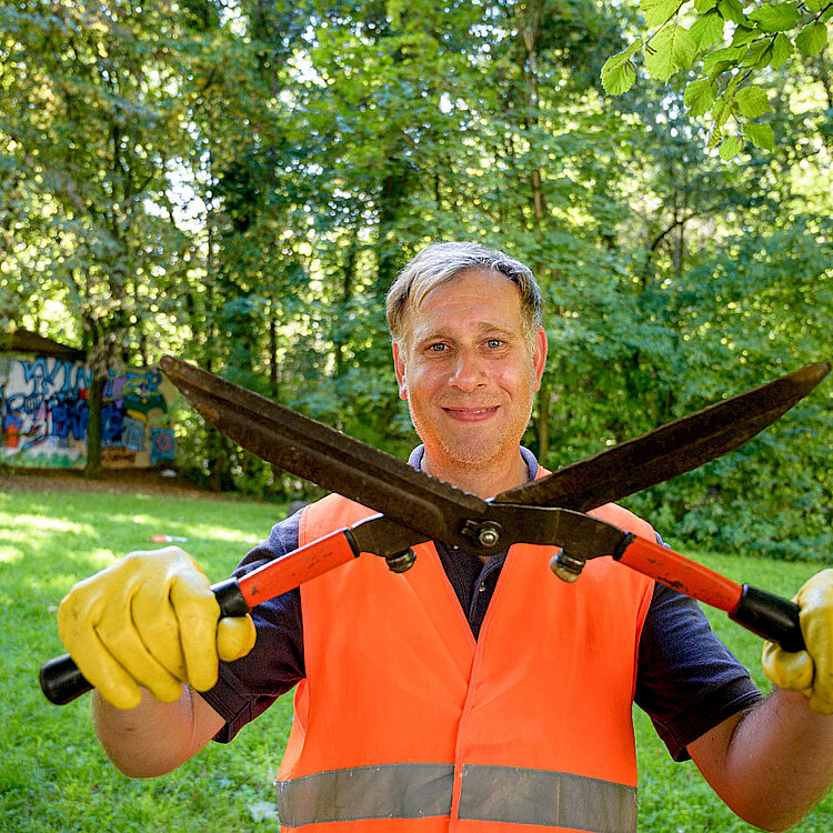 Ein Mann in oranger Warnweste und Handschuhen hält zwei Heckenscheren gekreuzt vor sich, umgeben von grünem Wald und Gras.