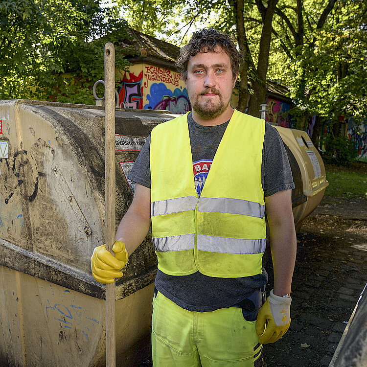 Ein Mann mit gelber Warnweste und Handschuhen steht vor großen Müllcontainern und hält einen langen Besenstiel, symbolisiert Müllabfuhr oder Reinigung im öffentlichen Raum.