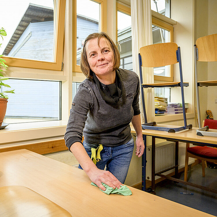 Frau reinigt in hellem Raum mit großen Fenstern einen Holztisch, zwei Stühle stehen auf einem anderen Tisch. Sie trägt grauen Pullover und Jeans und lächelt in die Kamera.