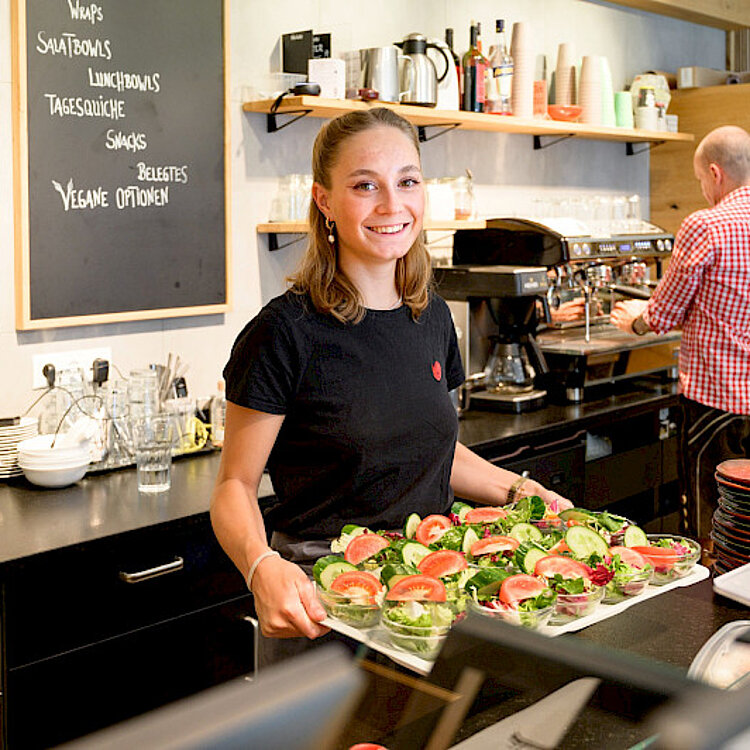 Eine junge Frau in einem Café trägt ein Tablett mit frisch zubereiteten Sandwiches und Salaten. Im Hintergrund bedient ein Mann eine Kaffeemaschine. Die Atmosphäre wirkt freundlich und einladend.