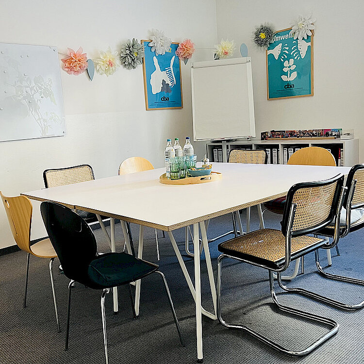 Konferenzraum mit einem großen, rechteckigen weißem Tisch, umgeben von verschiedenen Stühlen. Auf dem Tisch stehen Wasserflaschen und Gläser. An der Wand hängen zwei Plakate, eine Girlande aus Papierblumen und ein Flipchart. Der Raum wirkt hell und einladend.