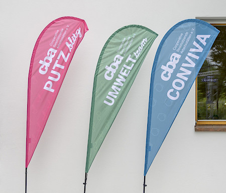 Drei bunte Werbefahnen lehnen an einer weißen Wand, beschriftet mit „cba PUTZ flink“ (pink), „cba UMWELT team“ (grün) und „cba CONVIVA“ (blau).