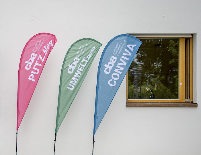 Drei bunte Werbefahnen vor einer weißen Wand mit Fenster, beschriftet mit „PUTZ blitz“, „UMWELT team“ und „CONVIVA“ in großen Buchstaben.