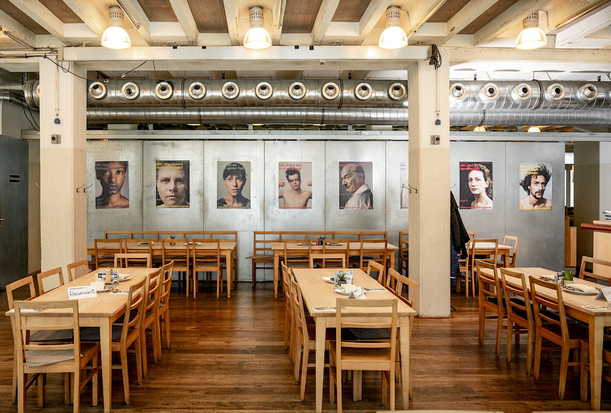 Blick in einen hellen, rustikalen Gastraum mit Holztischen und -stühlen, an der Wand hängen große Porträtfotos verschiedener Menschen.