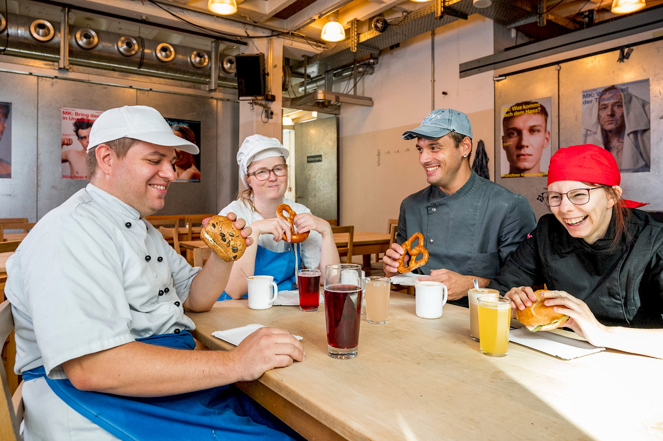 Vier Bäcker in Arbeitskleidung sitzen an einem Holztisch, essen Brezeln und andere Backwaren und trinken verschiedene Getränke, sie lachen und unterhalten sich freundlich.