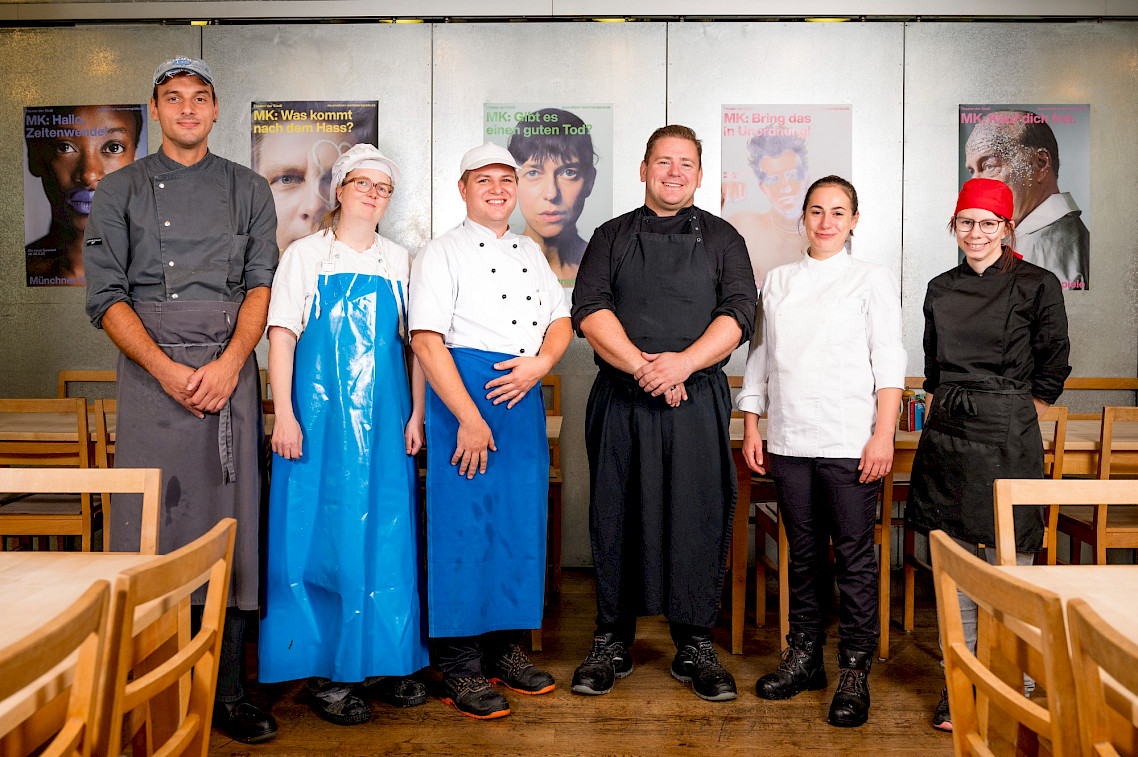 Sieben Personen in Küchen- und Kochkleidung stehen nebeneinander in einem Speisesaal. Sie wirken als Team von Küchenmitarbeitenden oder Köchen.