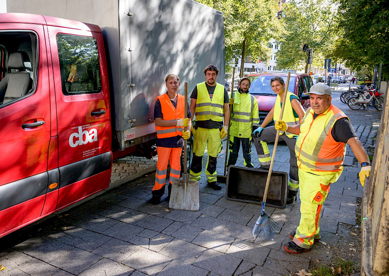 Fünf Straßenarbeiter in neongelber und oranger Warnkleidung stehen auf einem Bürgersteig neben einem roten Fahrzeug. Sie halten Besen und Schaufeln und blicken in die Kamera. Im Hintergrund sind Bäume und geparkte Fahrräder zu sehen.