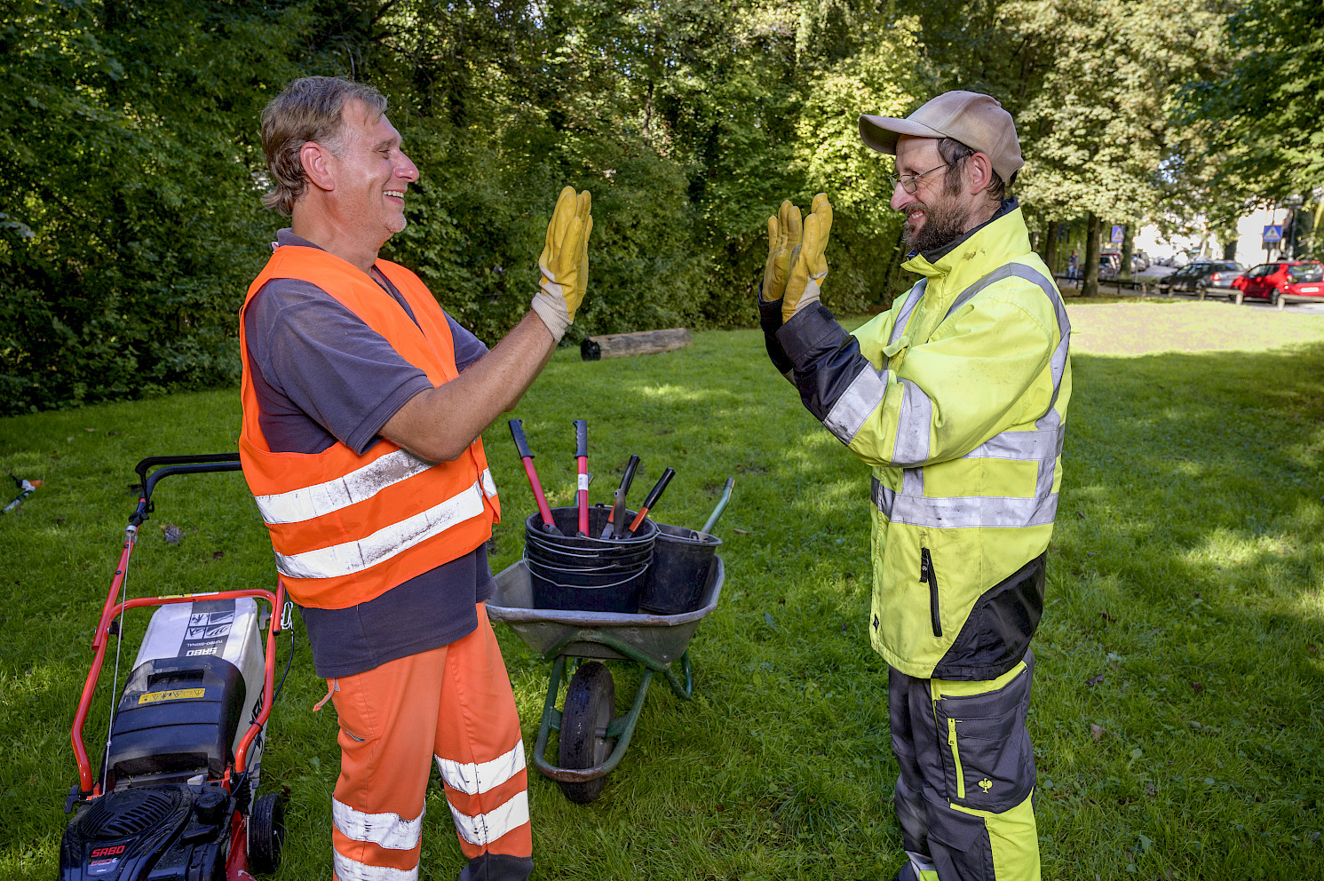 Zwei Männer in Arbeitskleidung stehen auf einer Wiese und geben sich freundschaftlich ein High-Five, neben ihnen stehen Gartenwerkzeuge und eine Schubkarre.