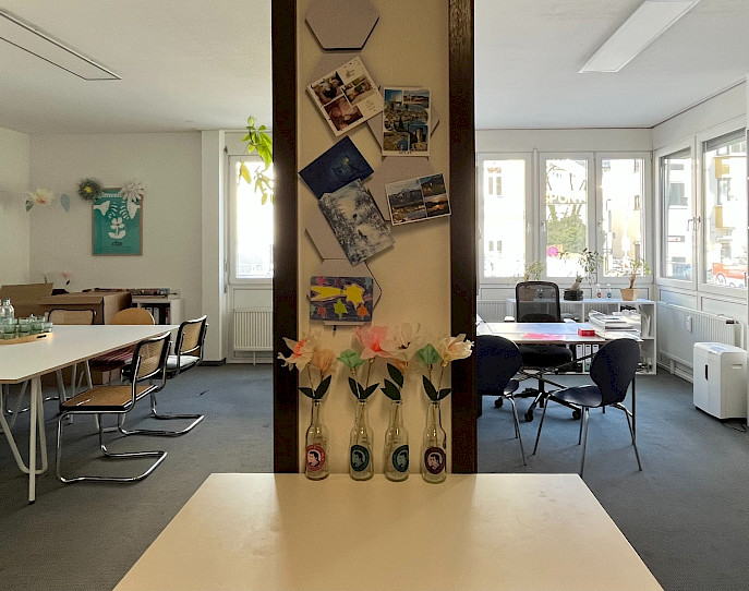 Ein heller, moderner Büroraum mit mehreren Tischen und Stühlen. Im Vordergrund stehen vier Glasflaschen mit Papierblumen und Postkarten an einer Wand, die den Raum in zwei Bereiche unterteilt. Viel natürliches Licht kommt durch große Fenster.
