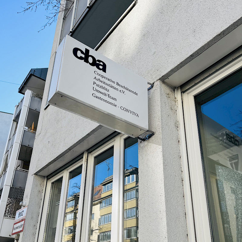 Außenaufnahme eines Gebäudes mit einem Schild, auf dem „cba Cooperative Beschäftigende Arbeitsstätten e.V. Publizistik Umwelt-Team Gastronomie CONVIVA“ steht.